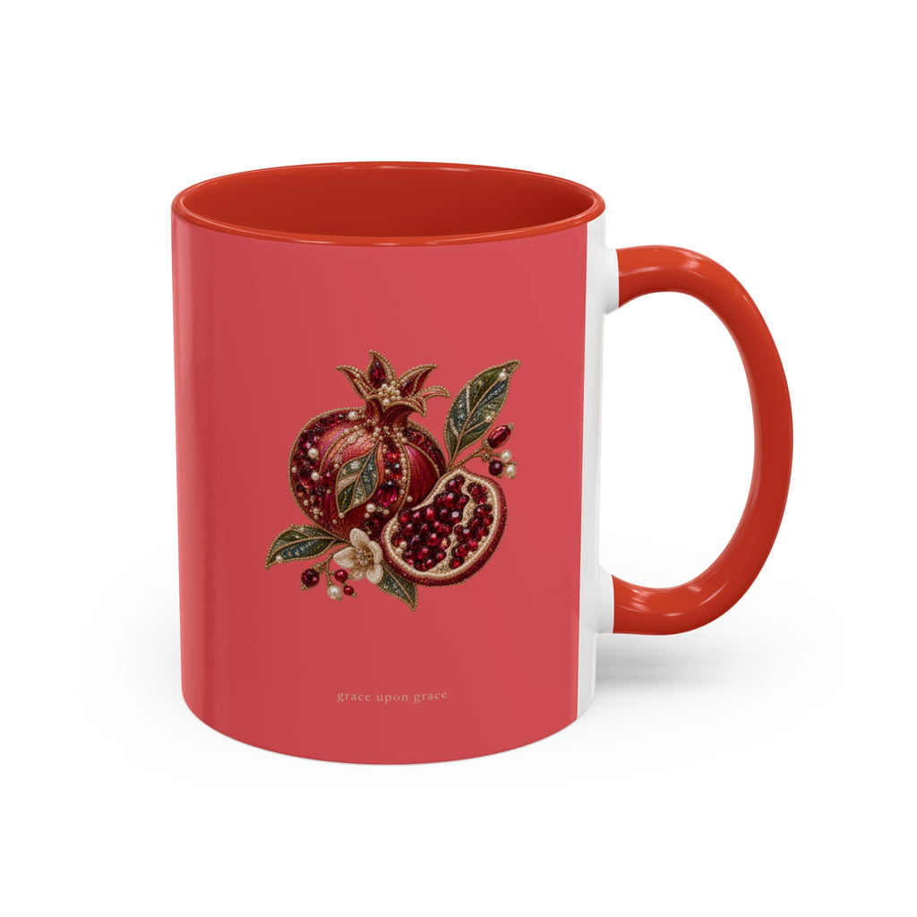 A ceramic mug with a coral red exterior, a white interior, and a handle matching the exterior. The front of the mug features a detailed illustration of a pomegranate, both whole and sliced open to reveal seeds, accompanied by green leaves and small flowers.