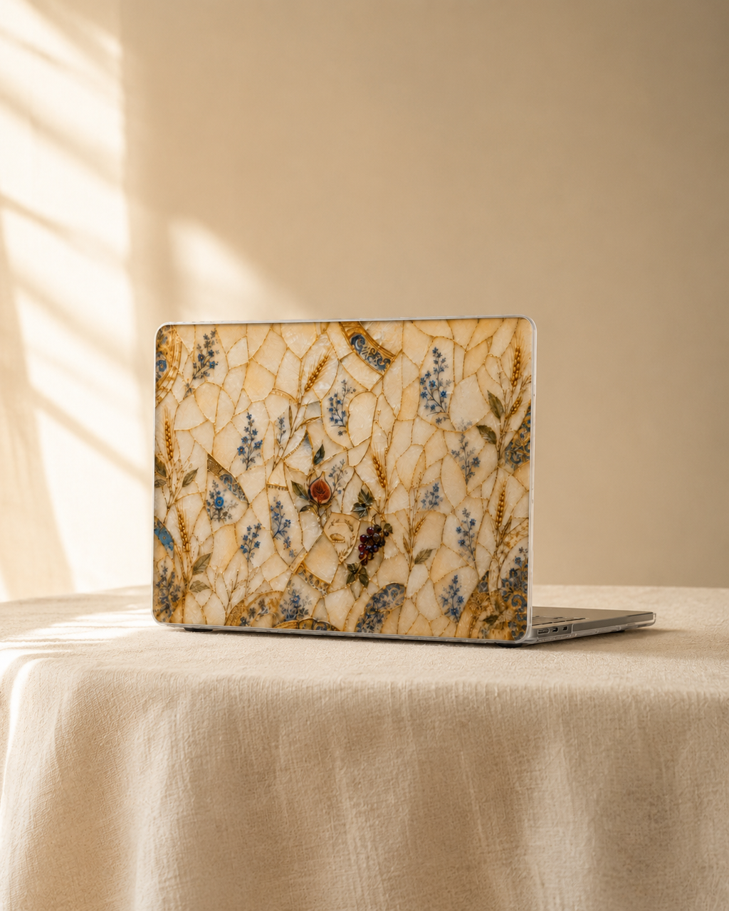 A closed laptop with a decorative beige mosaic-style skin featuring blue and gold botanical patterns, resting on a cream tablecloth in warm natural light.