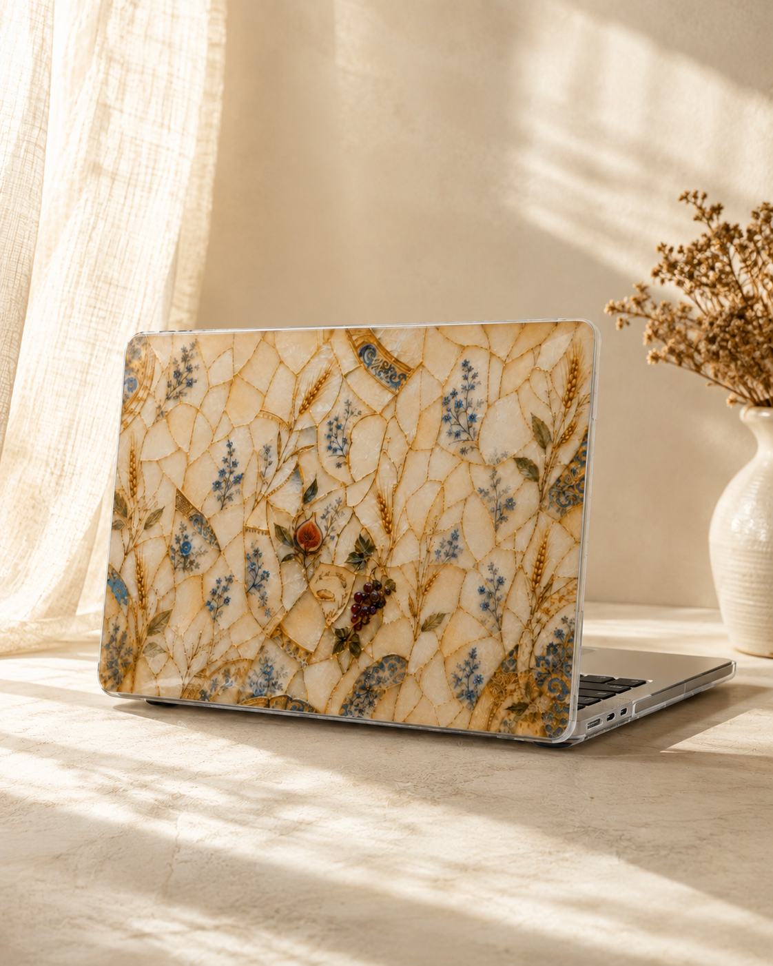 A laptop with a decorative mosaic-patterned skin in beige, gold, and blue botanical designs, resting on a table in a warm, sunlit room next to a vase of dried flowers.