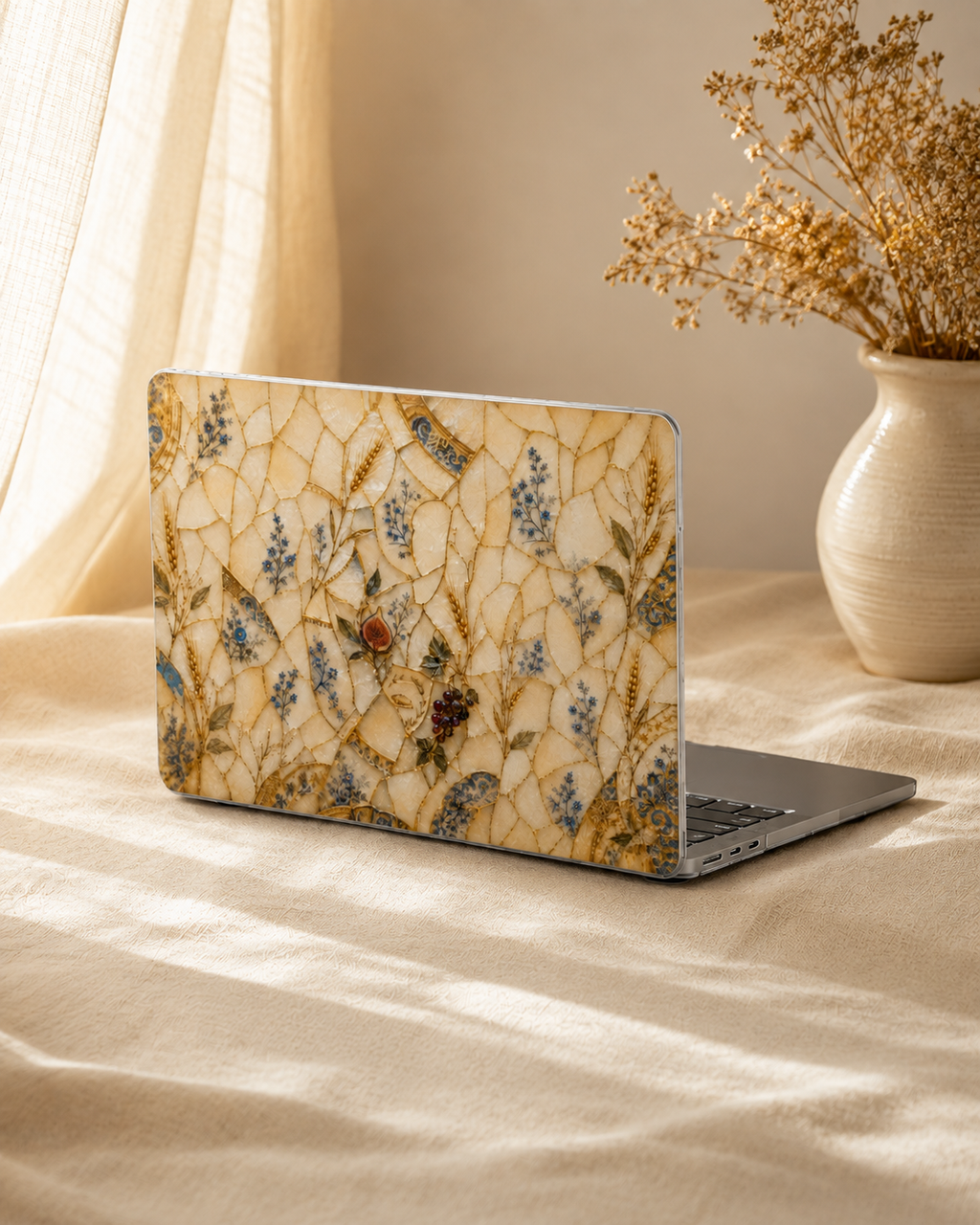 A laptop with a decorative floral and insect patterned skin on the back, resting on a beige fabric surface next to a vase of dried flowers.